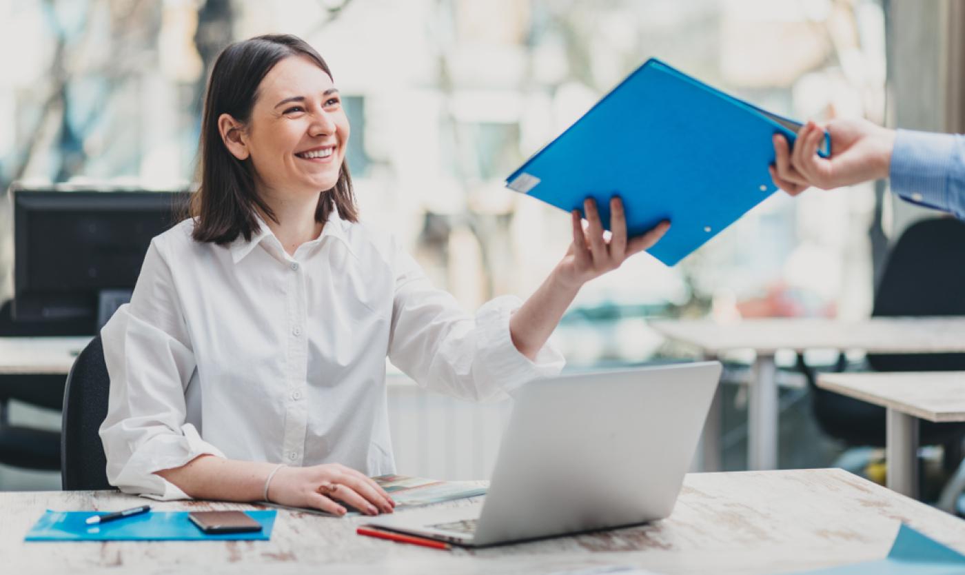 2022_Financiële ondersteuning_Samen Innoveren_Shutterstock_1040377084 Persoon die blauwe map overhandig aan persoon achter een laptop