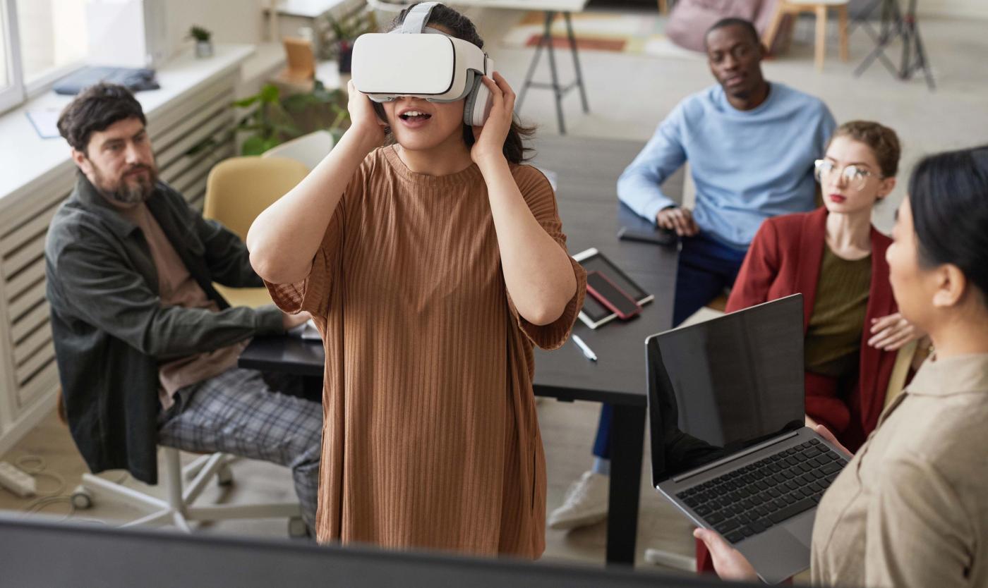 2022_Innoveren_VR_Shutterstock_1945476319 Iemand kijkt door een VR bril groepje mesen staan er rond en kijken naar de persoon met VR-bril