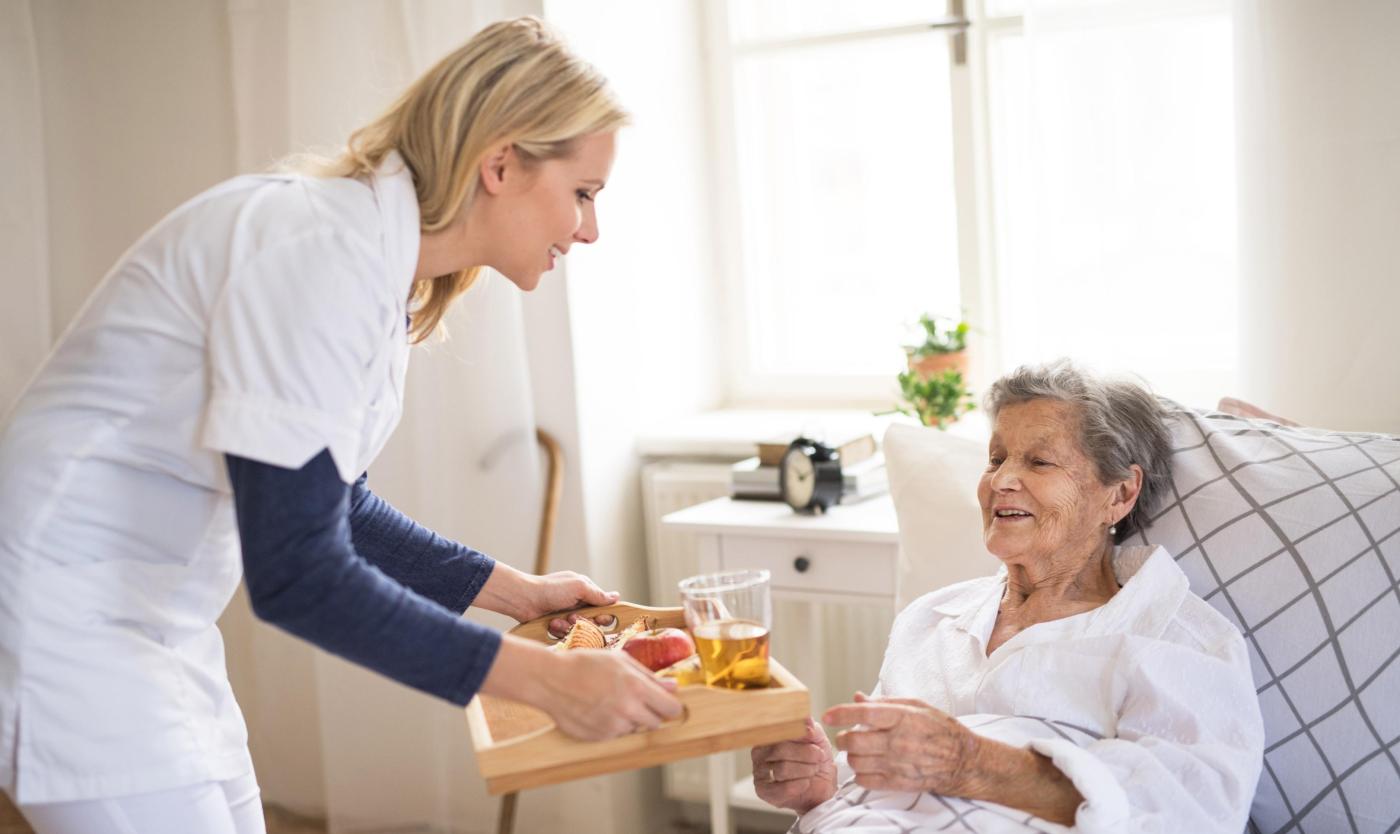 2022_Een gezondheidsbezoeker die ontbijt brengt naar een zieke oudere vrouw die thuis in bed ligt_Slaapkamer_S_1228707130