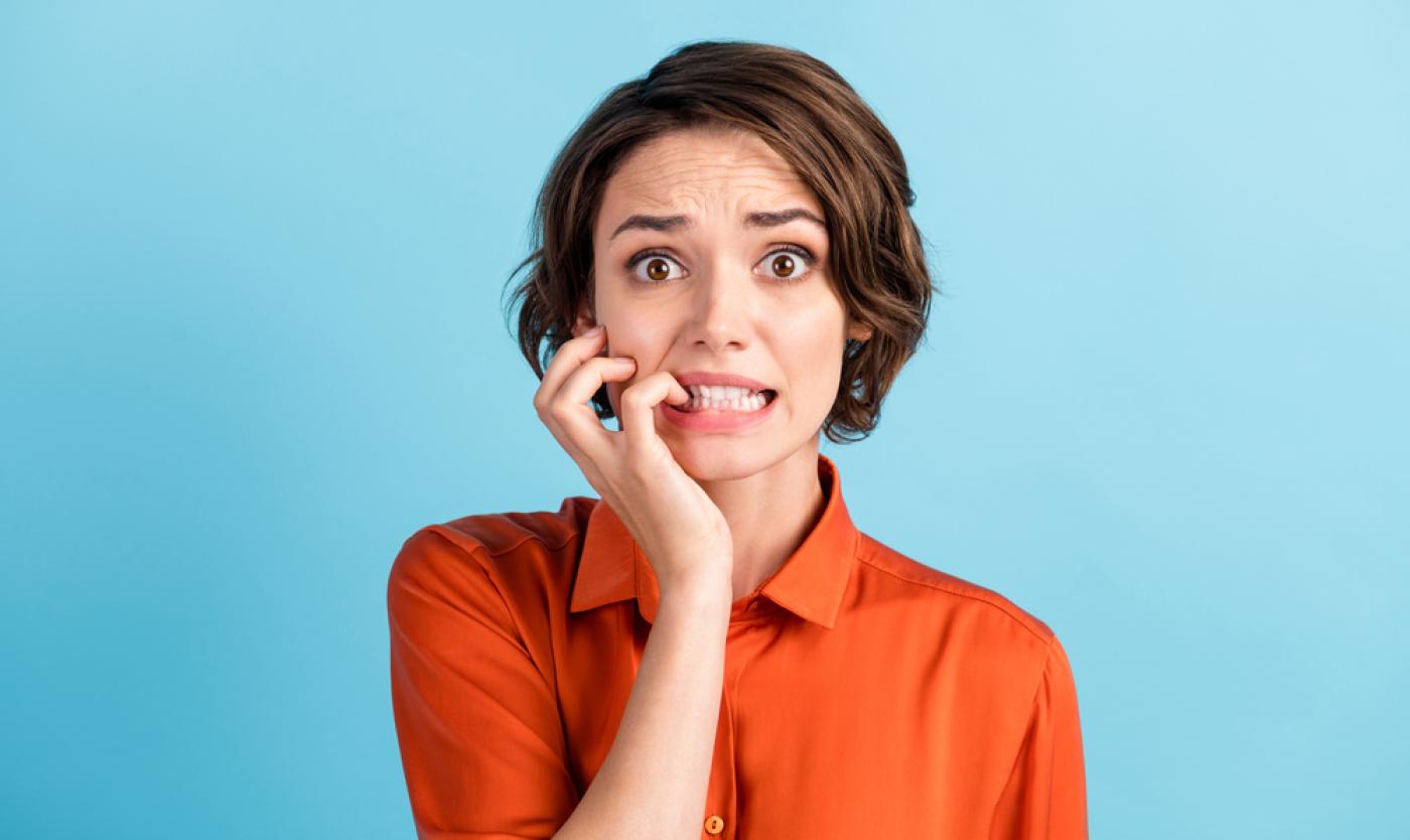 Vrouw in oranje bloes die op haar nagels bijt. Ze kijkt angstig. 